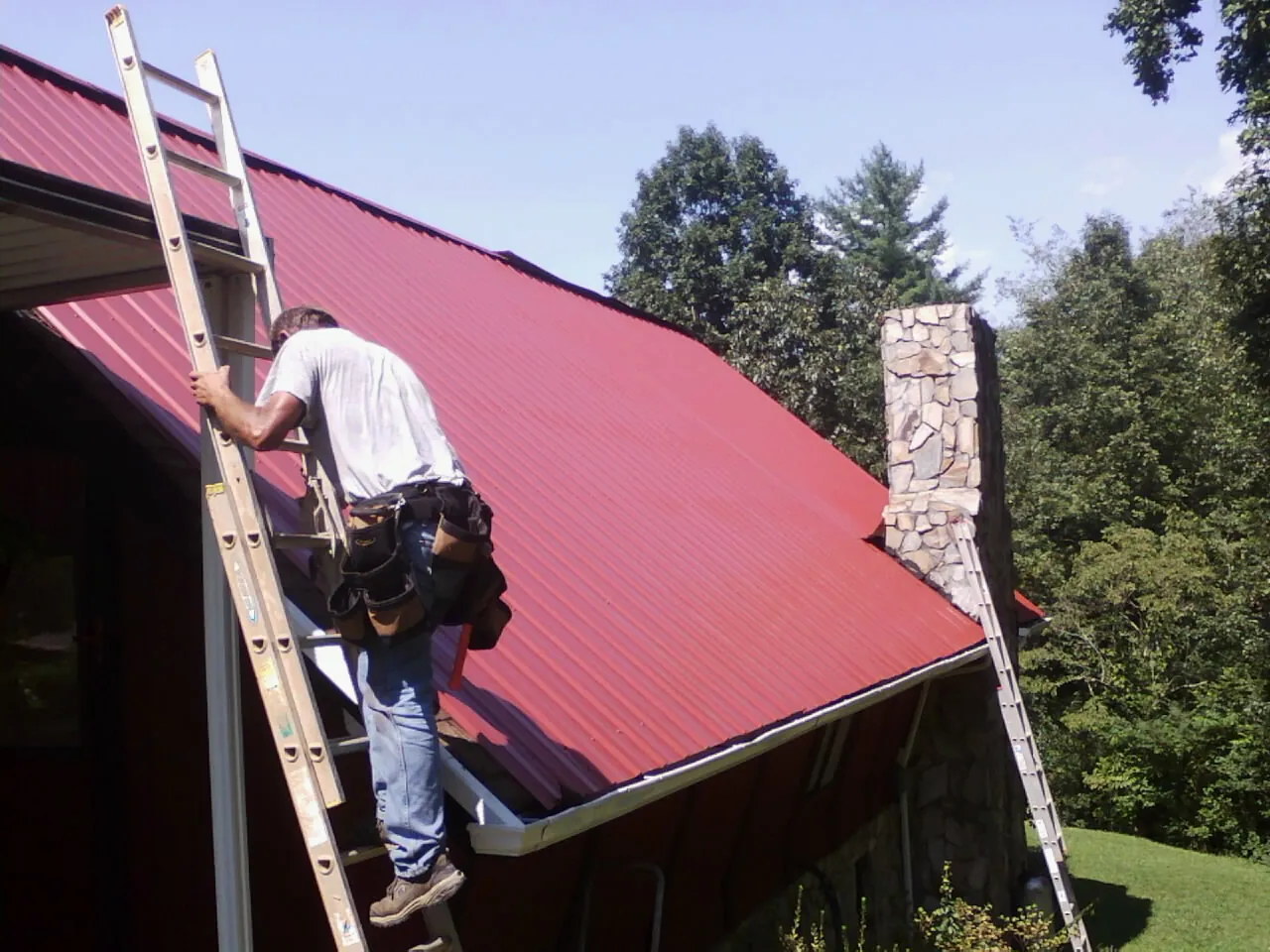 Roofer on a ladder beside a red metal roof and stone chimney — Soffit & Fascia Repair by Roofing Brothers in Alkol